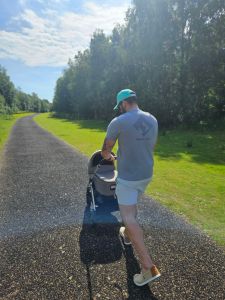 A man pushing a baby stroller along a tree-lined pathway on a sunny day.